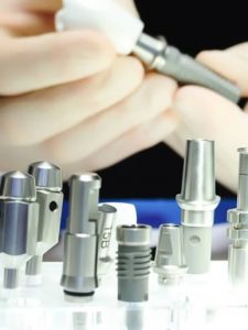 Implantate Pforzheim: Nahaufnahme mehrerer metallischer Zahnimplantat-Komponenten, die auf einer weißen Oberfläche angeordnet sind. Im Hintergrund montieren behandschuhte Hände präzise ein Implantatteil.